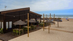 A beachside restaurant with outdoor seating, a wooden deck, and thatched umbrellas.