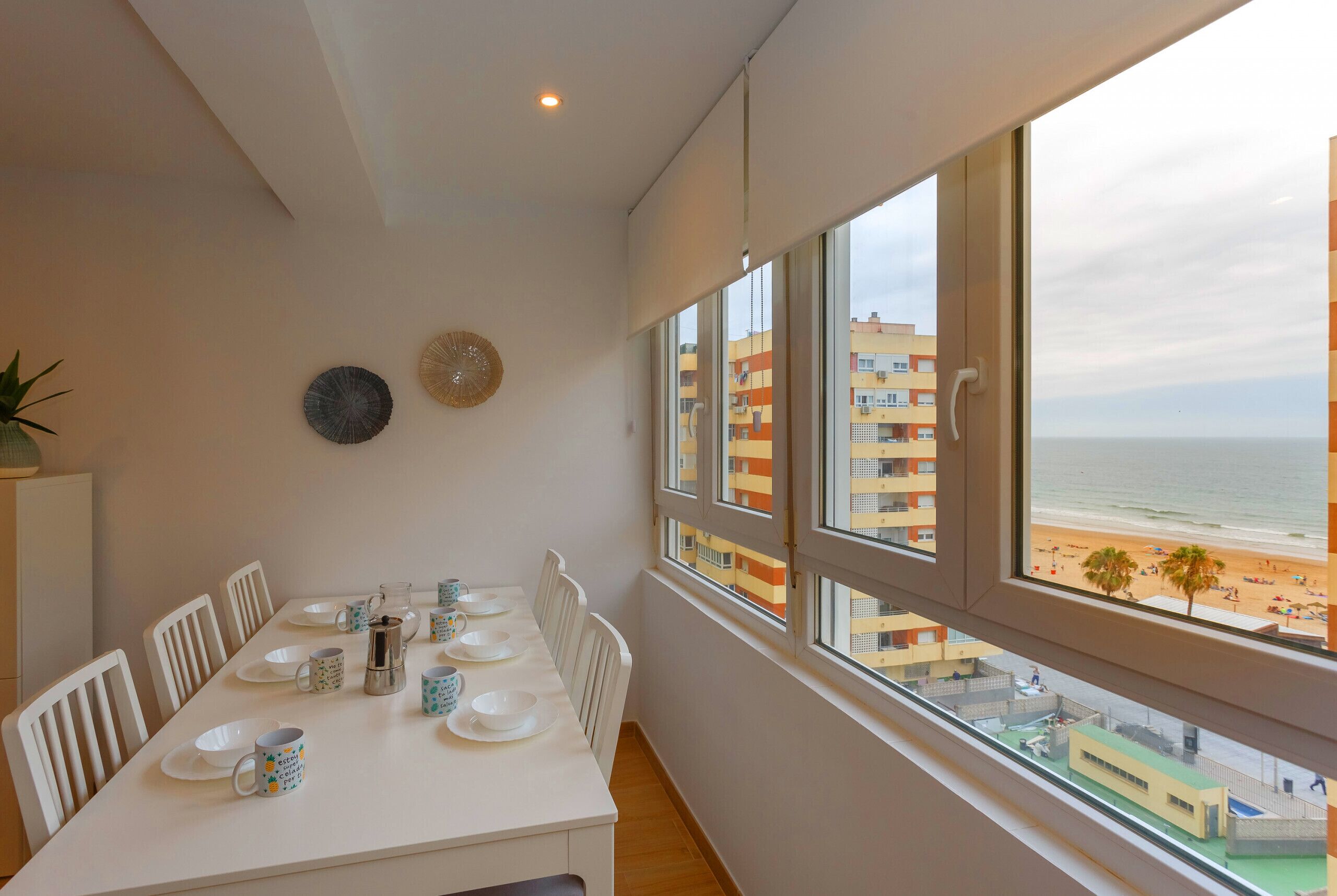 A dining area with a table set for four, a coffee pot, and plates. There are chairs around the table, and a view of a beach and buildings through large windows.