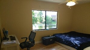 Un dormitorio con una cama, un escritorio con una silla, una maleta y una ventana con vistas a árboles y una casa.