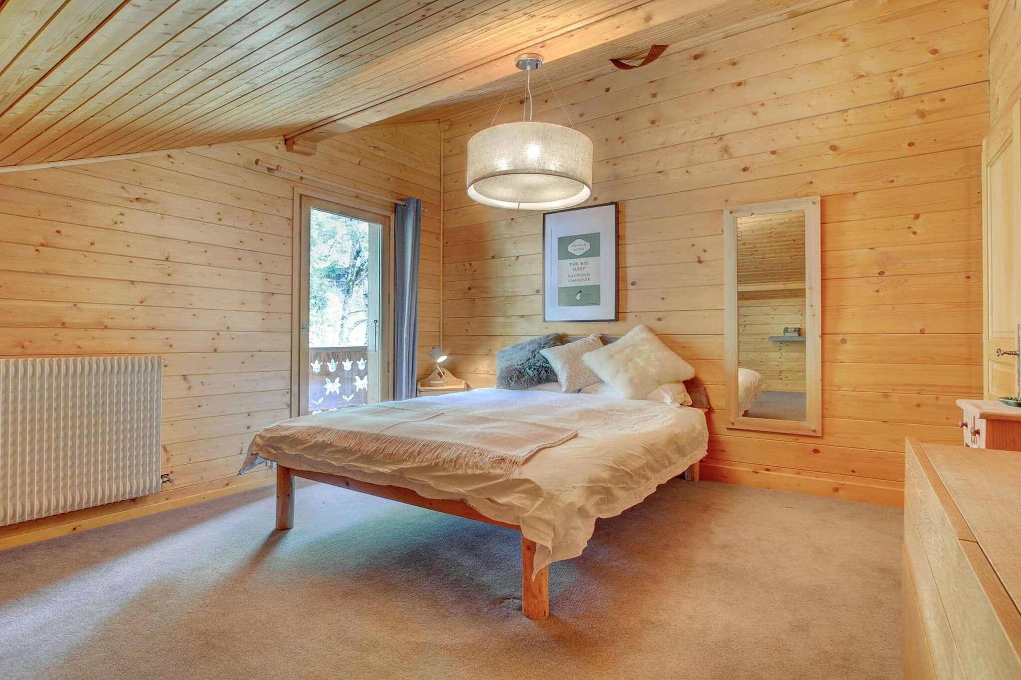 A wooden cabin bedroom with a bed, a hanging light fixture, a framed picture on the wall, and a window with a view of trees.
