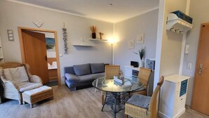 Ein Wohnzimmer mit einem Glastisch, Korbstühlen, einem grauen Sofa und einer kleinen Kommode.