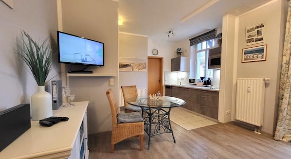 A modern living room with a flat-screen TV, a dining table with chairs, a small kitchen area with a microwave, and a radiator.
