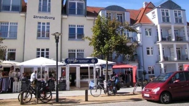 Straßenansicht mit Gebäuden, auf denen 'Strandburg' und 'FASCA' steht. Vor einem Gebäude stehen Fahrräder, und ein rotes Auto ist auf der Straße sichtbar.