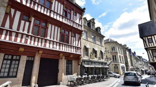 A street with traditional half-timbered buildings, a café with outdoor seating, and parked cars. - Martin pecheur - Ferme des Herils (Port-en-Bessin-Huppain)