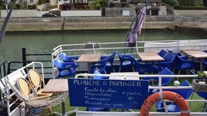 A boat with tables and chairs, a sign listing food items, and a view of houses along the water. - Martin pecheur - Ferme des Herils (Port-en-Bessin-Huppain)