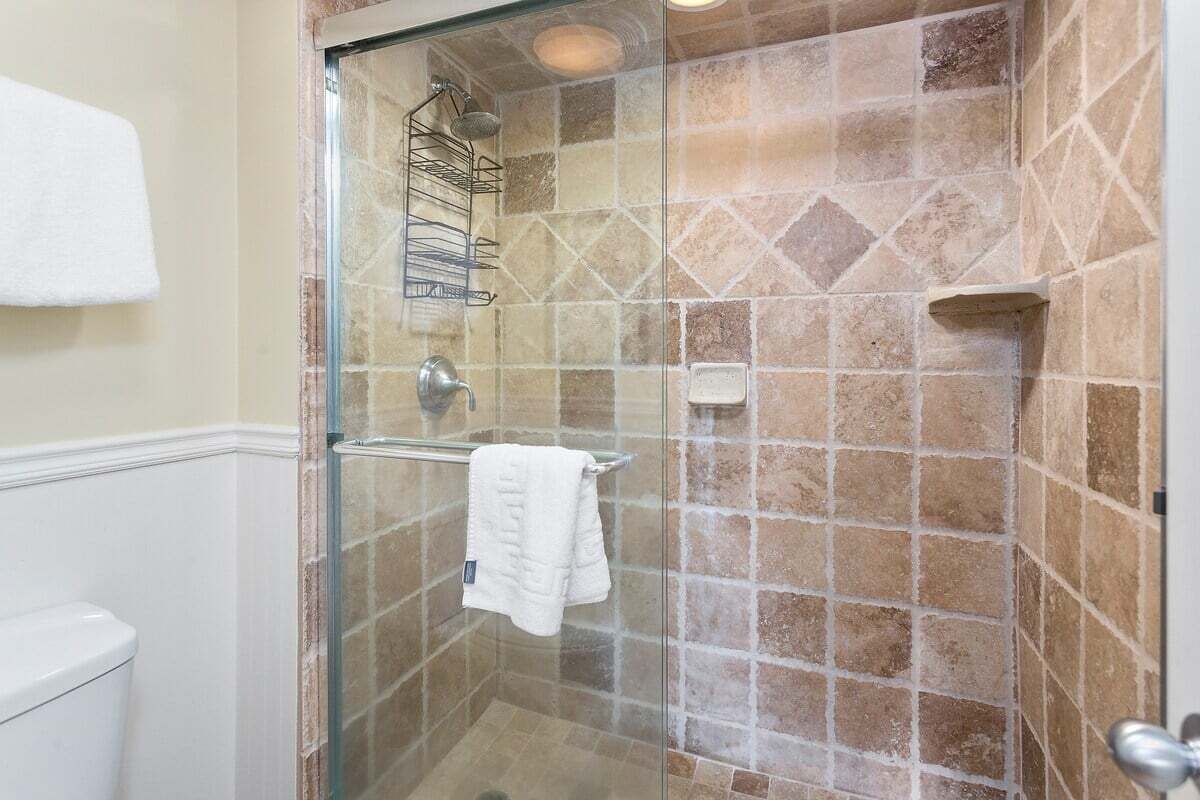 A glass-enclosed shower with a tiled wall, a white towel hanging on a rack, and a small shelf.