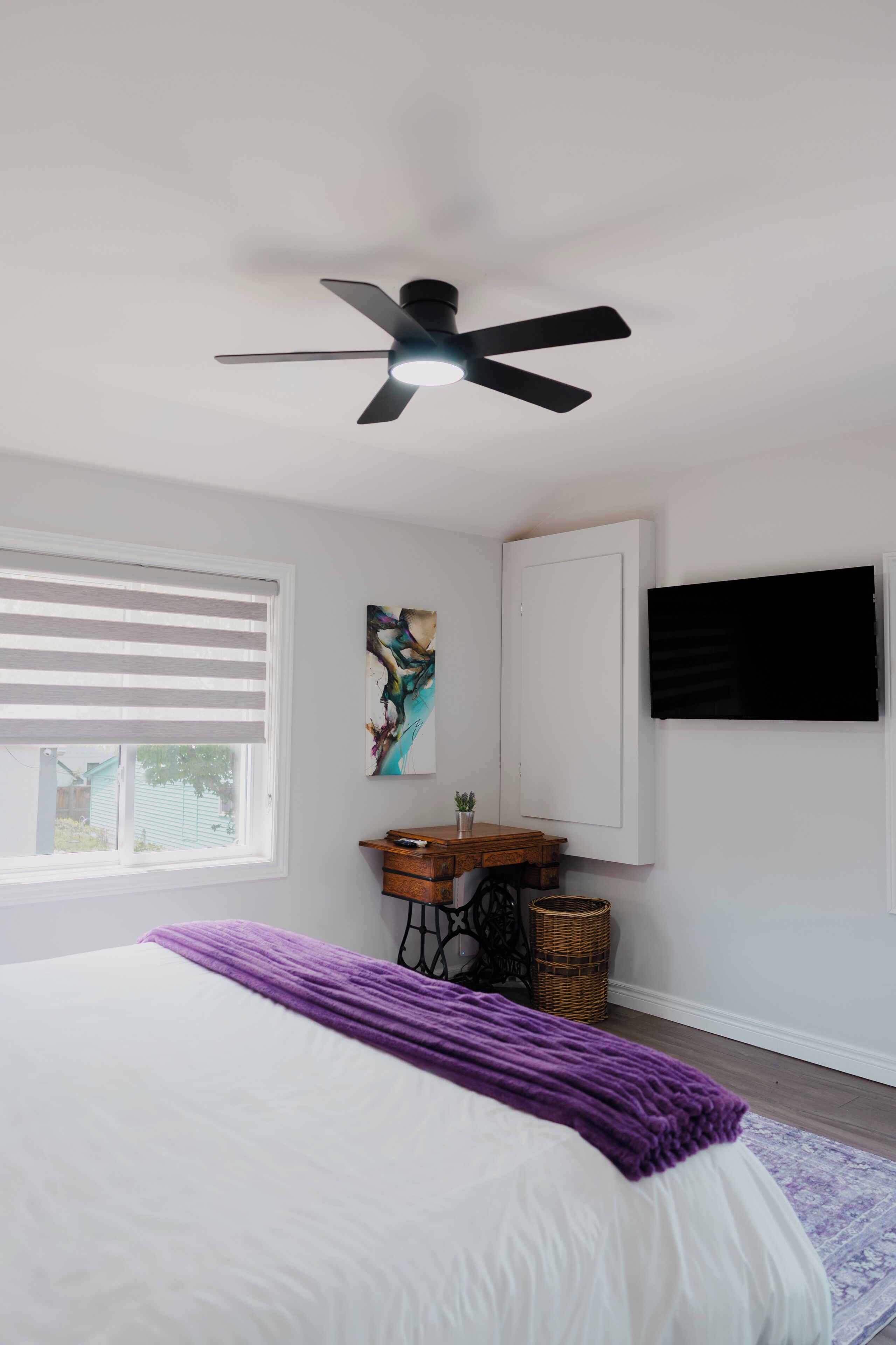 Een slaapkamer met een bed, een plafondventilator, een raam met jaloezieën, een schilderij, een televisie, een klein tafeltje en een rieten mand.
