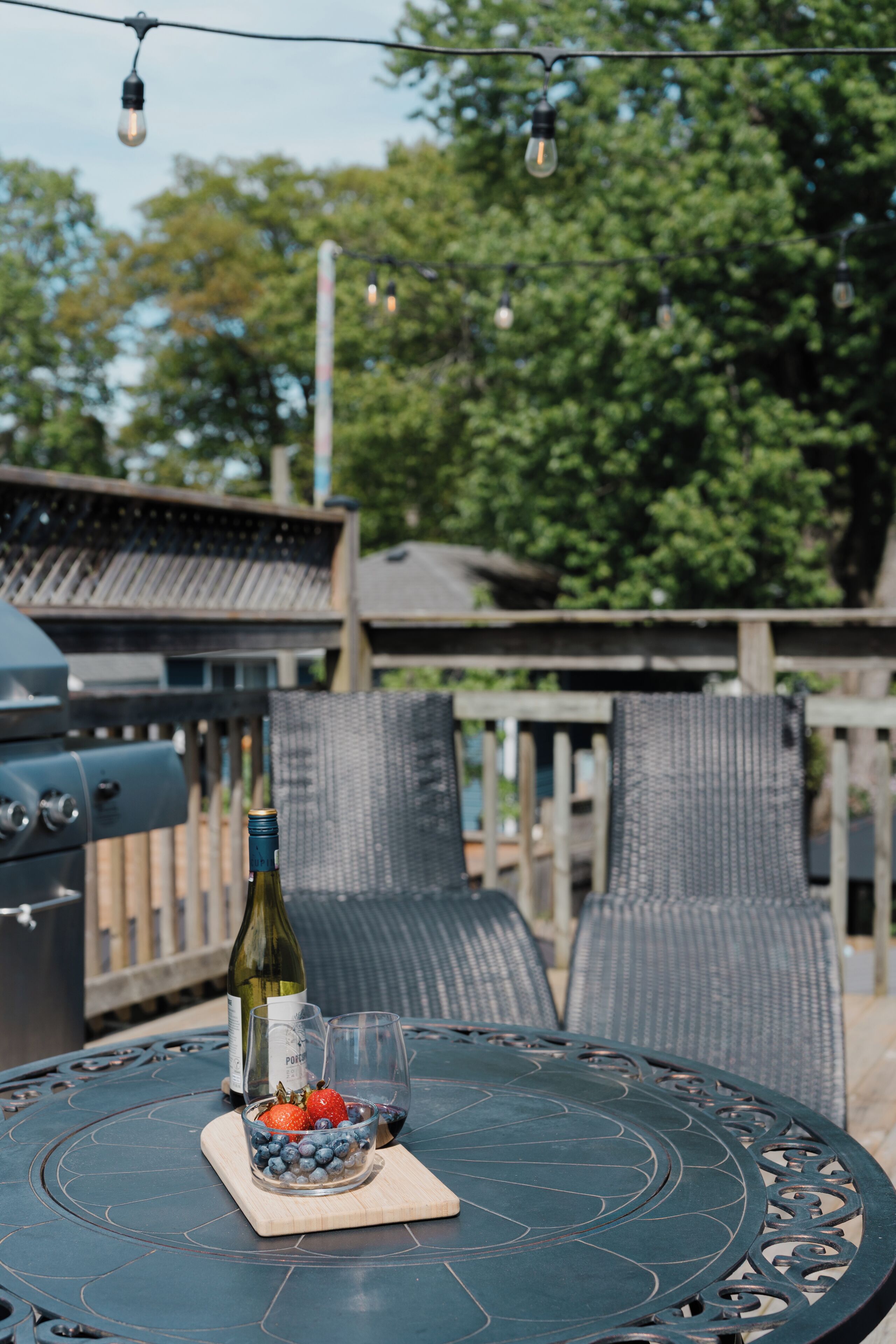 A round outdoor table with a bottle and glasses, a bowl of fruit, and a grill in the background.