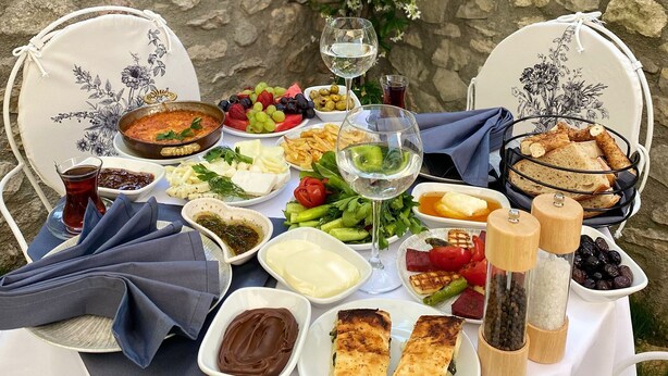 A table set with a variety of dishes, including a bowl of soup, fruits, cheese, bread, and condiments, against a stone wall with greenery. - Ahşena Bağ Badeli Konak Safranbolu (Safranbolu)