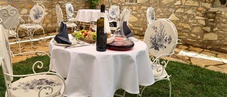 Mesa redonda dispuesta para una comida, con mantel blanco, rodeada de sillas ornamentadas con motivos florales, sobre un fondo de pared de piedra.