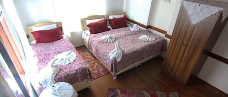A room with two beds, each with a red pillow and a white towel. There is a wooden wardrobe and a patterned rug on the floor.