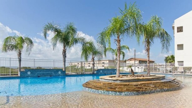 A swimming pool with palm trees, a tiled deck, and a white building in the background. - Bay Front: Gradual-Entry Pool: Boat Slip (South Padre Island)