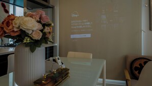 Ein modernes Zimmer mit Glastisch, Blumenvase, Projektorbildschirm mit Text und einem Sessel mit Kissen.