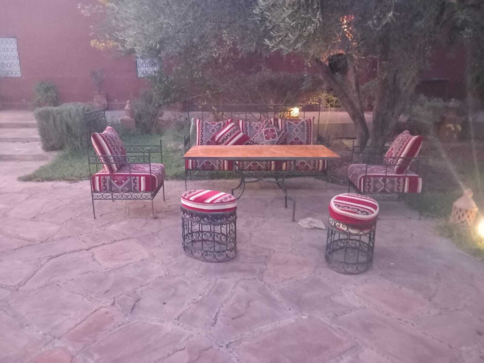 An outdoor patio with a wooden table and four patterned chairs, surrounded by stone flooring and greenery.