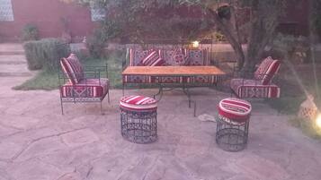 An outdoor patio with a wooden table and four patterned chairs, surrounded by stone flooring and greenery.