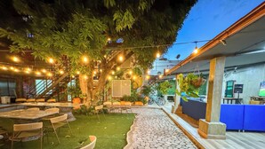 Un espace extérieur aménagé en terrasse, avec des tables et des chaises, un chemin en pierre et un arbre décoré de guirlandes lumineuses.