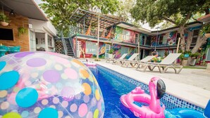 Une piscine avec des jouets gonflables aux couleurs vives, un ciel dégagé et un bâtiment orné d’une fresque colorée en arrière-plan.