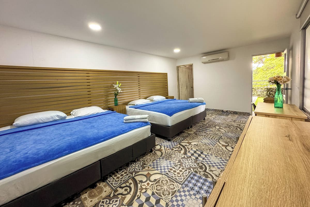 A hotel room with two beds, a wooden headboard, a patterned floor, a small table with a vase, and a window with a view of greenery.