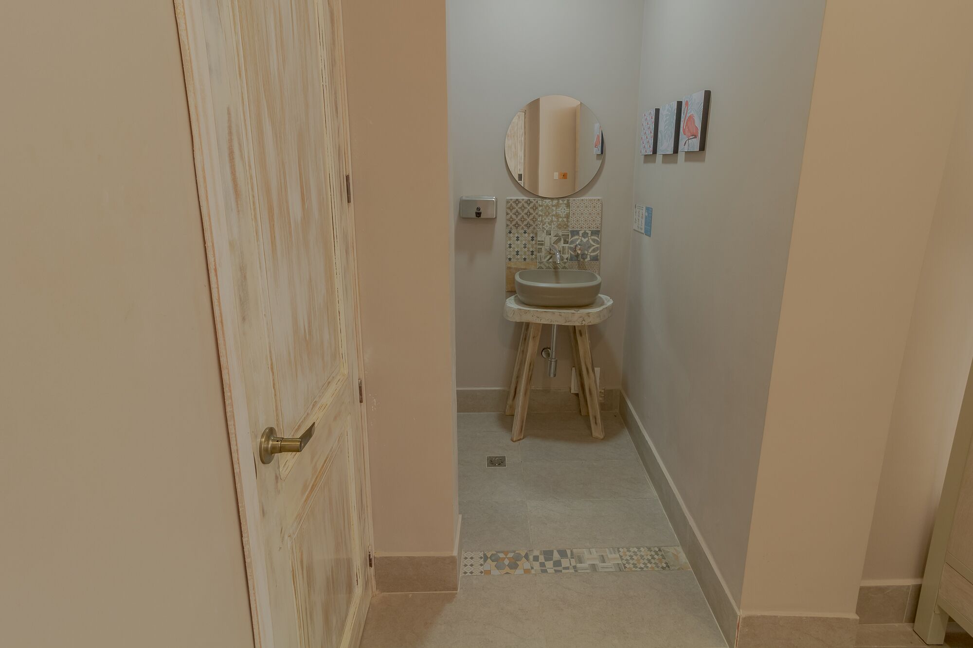 Um banheiro com porta de madeira, pia em pedestal, espelho e azulejos estampados na parede.