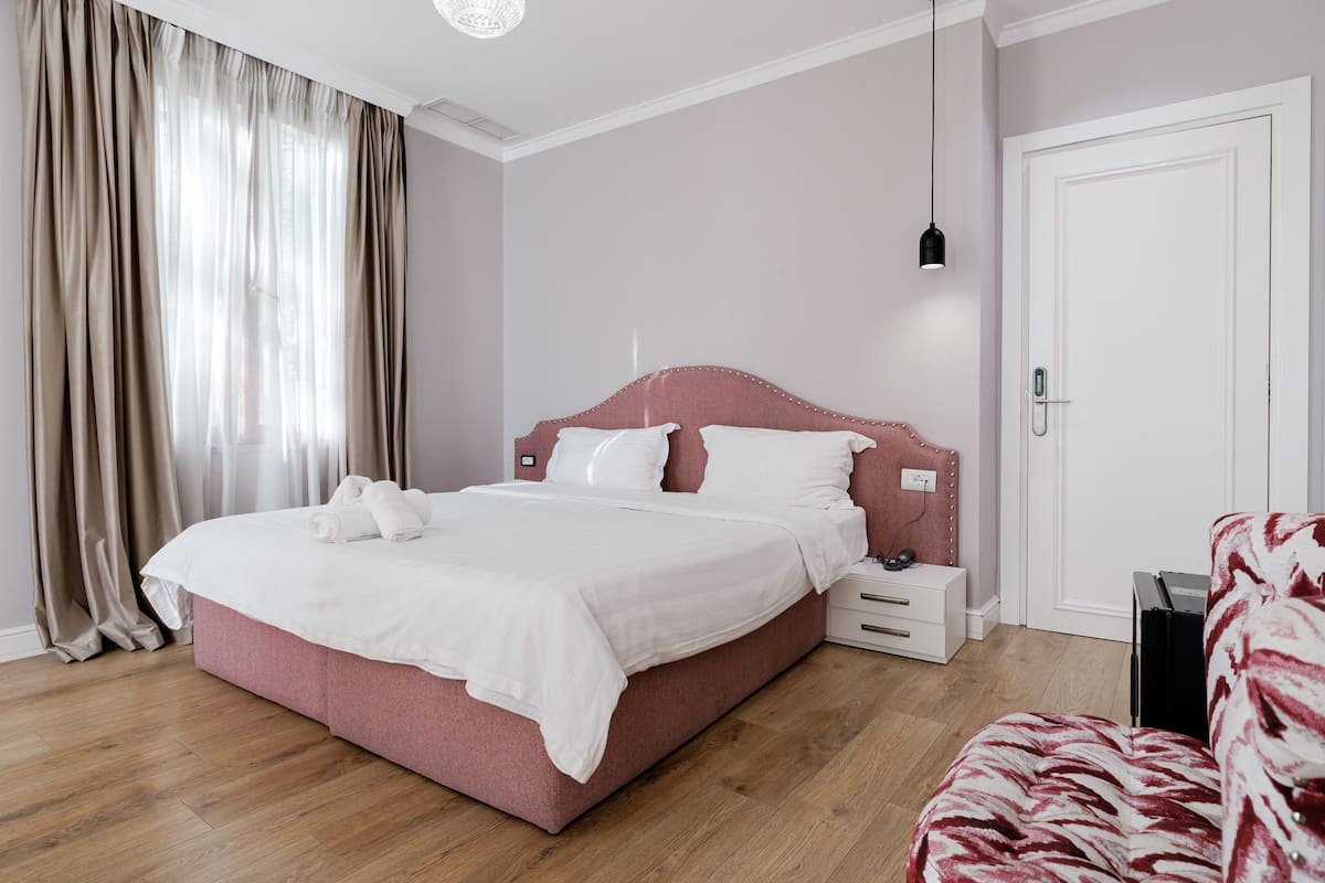 Ein ordentlich bezogenes Bett mit weißen Leinentüchern, ein Nachttisch mit Lampe sowie ein roter, gepolsterter Sessel.