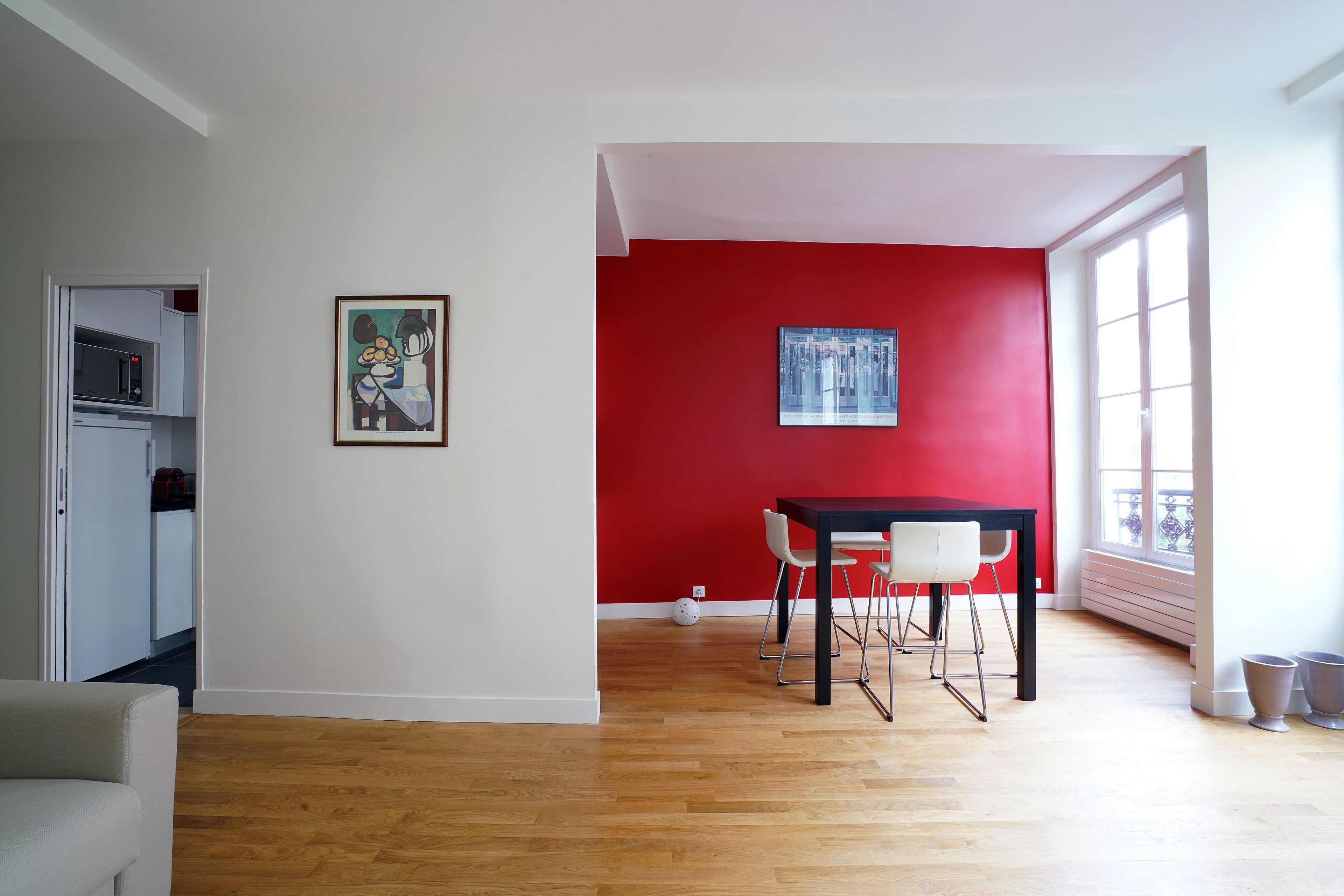 A room with a dining table, chairs, a red accent wall, and a window.