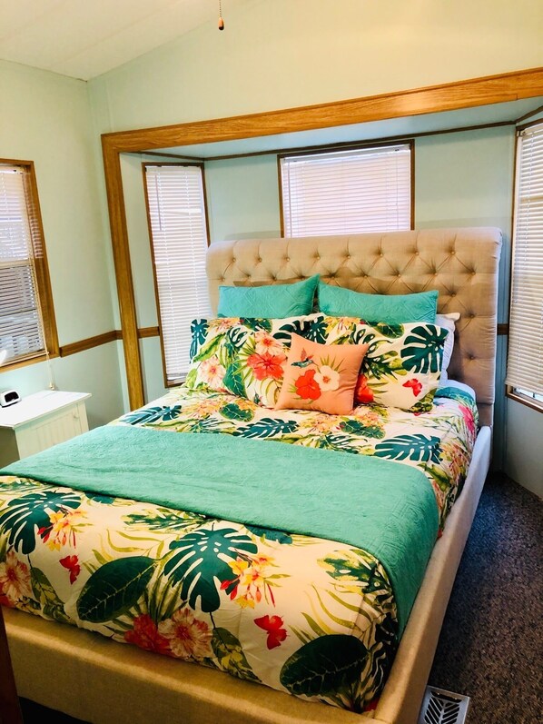 Una cama con una colcha estampada y un cabecero capitonado, flanqueada por ventanas con persianas.