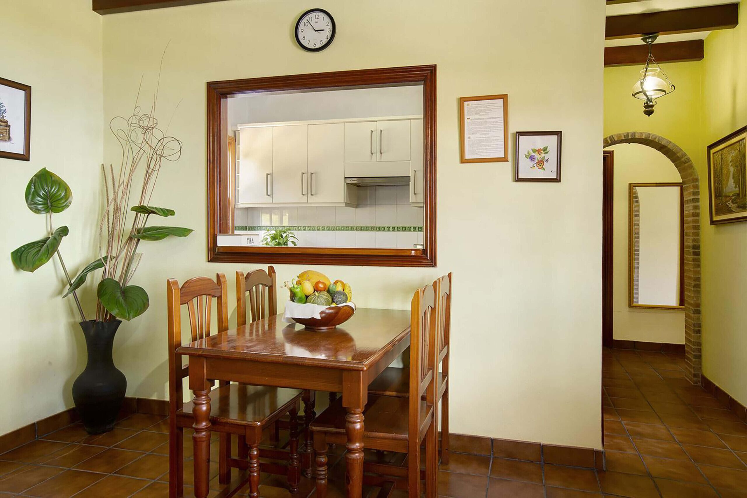 Un comedor con una mesa y sillas de madera, un florero con plantas y una ventana con vista a una cocina.