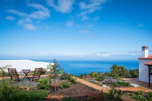 Une terrasse avec des chaises et une table, un jardin avec différentes plantes et une vue sur l’océan.