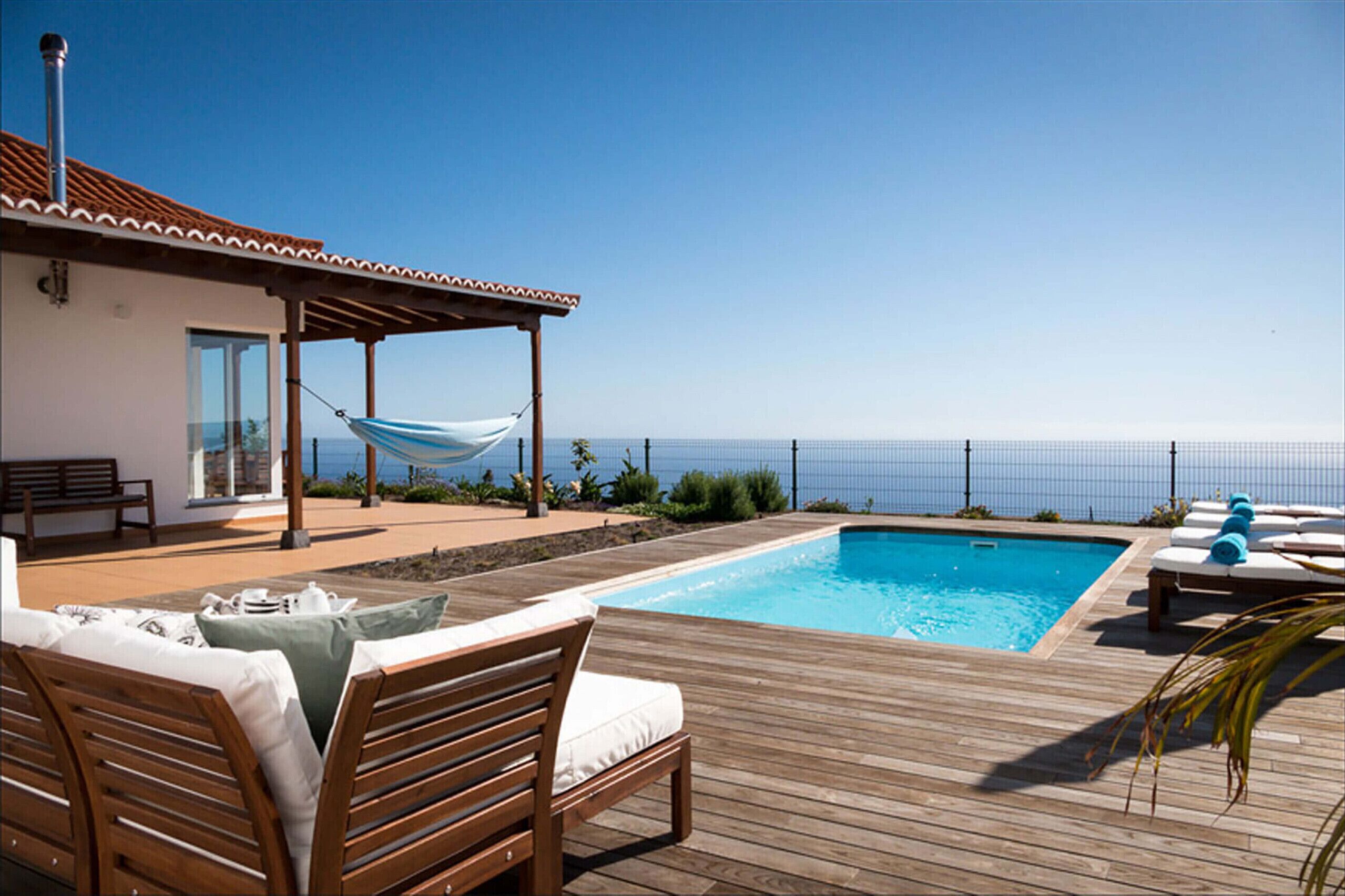Una terraza de madera con piscina, sillas de descanso, una hamaca y vista al océano.