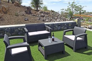 Outdoor wicker furniture with cushions, a wine bottle, and a glass on a square table, set against a backdrop of artificial grass and a rocky hillside. - Casa La Florida en Puntagorda (Tijarafe)