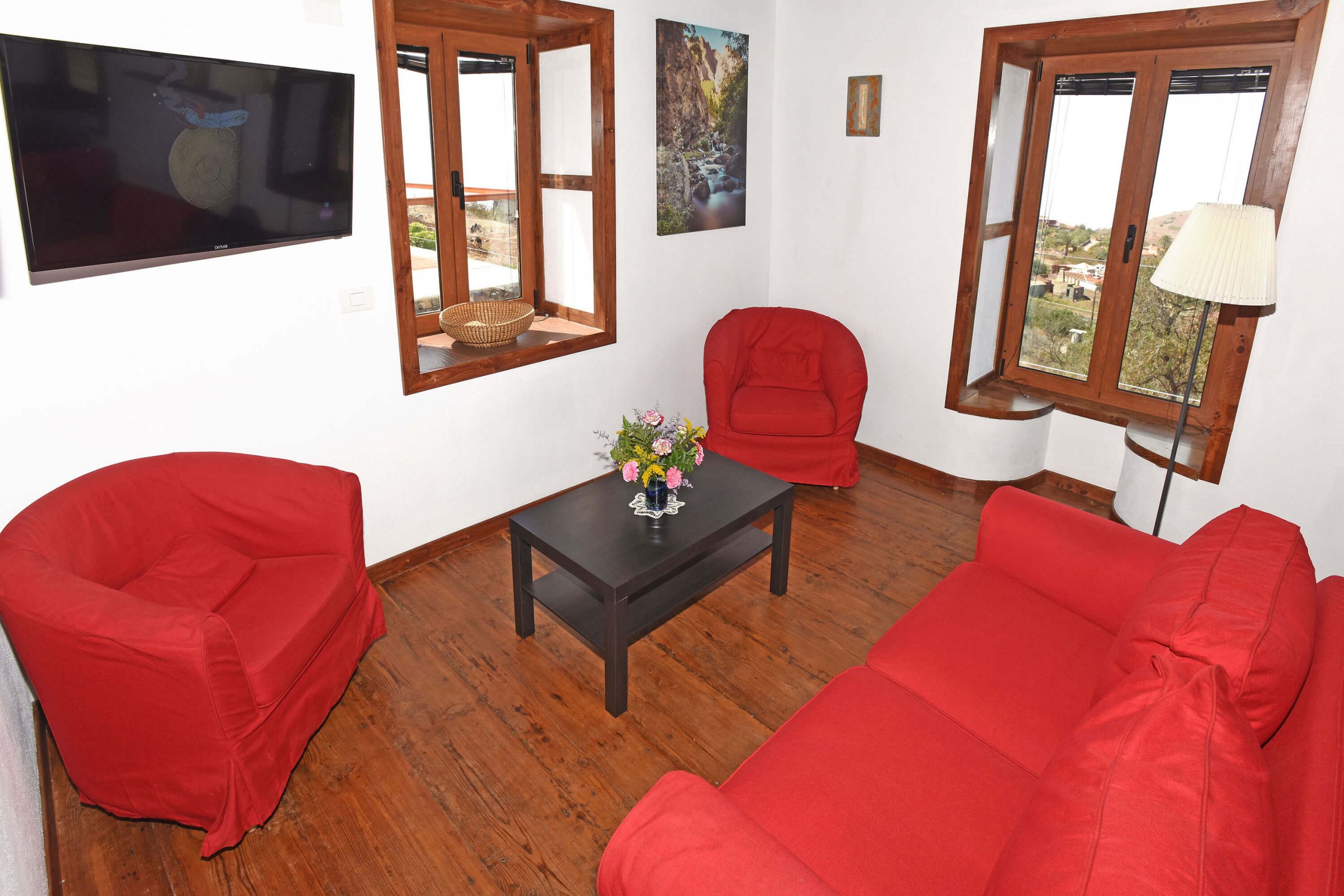 Un salon avec un canapé rouge, un fauteuil rouge, une petite table sur laquelle est posé un vase, et un téléviseur fixé au mur.