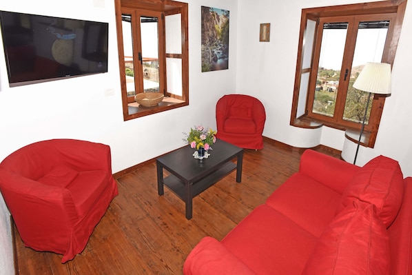 A living room with a red sofa, a red armchair, a small table with a vase, and a TV mounted on the wall.