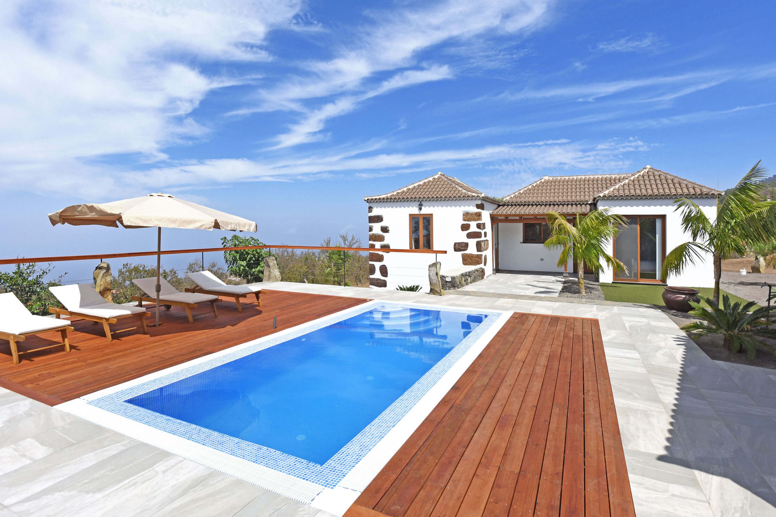 A swimming pool with wooden deck, white stone walls, and a clear blue sky.