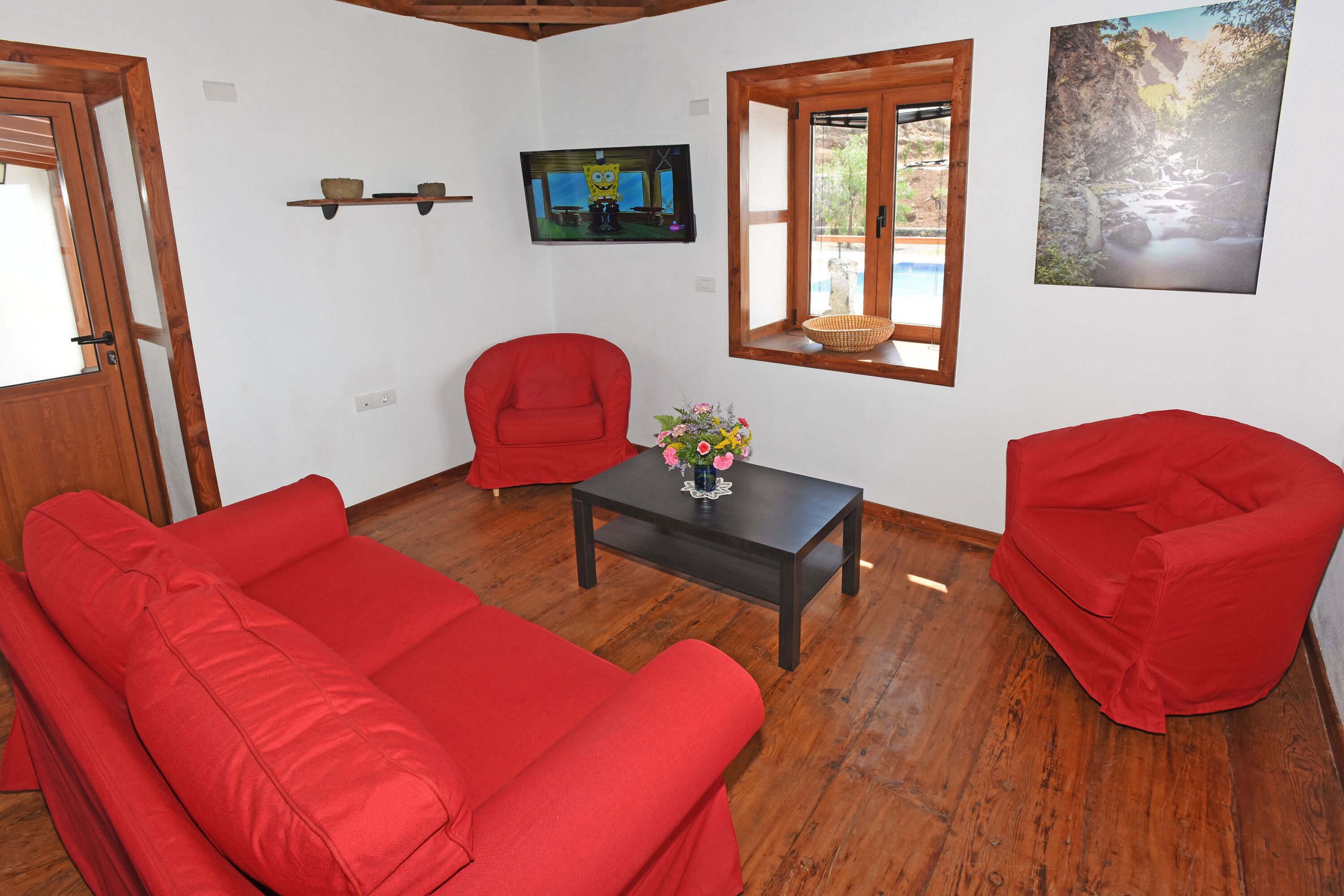 Una habitación con piso de madera, sofás rojos, una mesita con un florero y un televisor en la pared, además de una ventana con vistas a un paisaje rocoso.