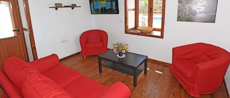 Une pièce avec un plancher en bois, des canapés rouges, une petite table sur laquelle est posé un vase de fleurs, un téléviseur fixé au mur et une fenêtre donnant sur un paysage rocheux.