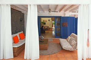 Une pièce avec un mur en pierre, un canapé en osier avec des coussins orange, un canapé à motifs et un ventilateur de plafond.