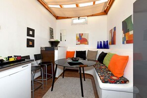 Ein kompakter Wohnraum mit einer Küchenzeile, einem runden Esstisch, einer Couch mit bunten Kissen und abstrakter Kunst an den Wänden.