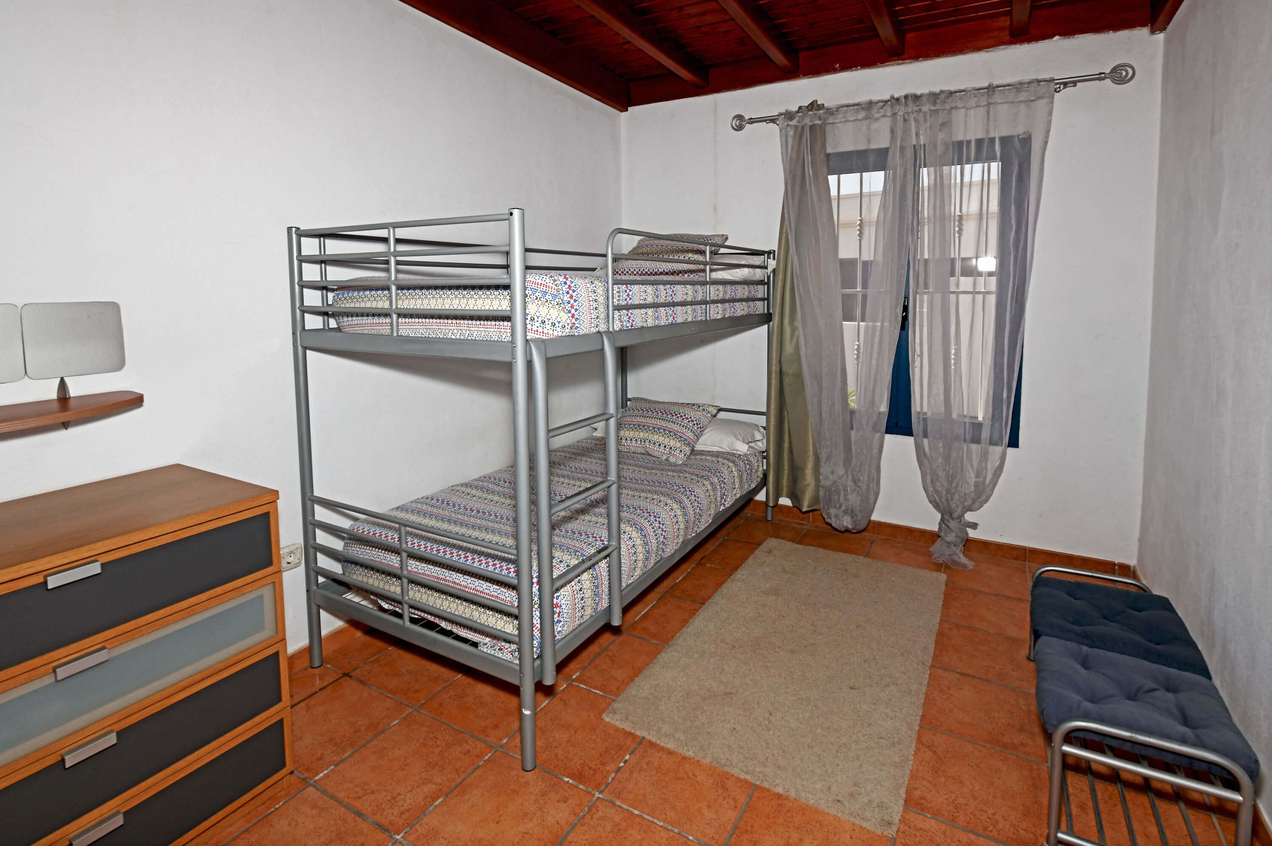 A bunk bed room with a desk and drawers, a window with curtains, and a small bench.