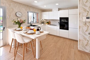 A modern kitchen with white cabinetry, a dining area with a table set for breakfast, and a clock on the wall. - Salobre Vista Golf 16 (Maspalomas)