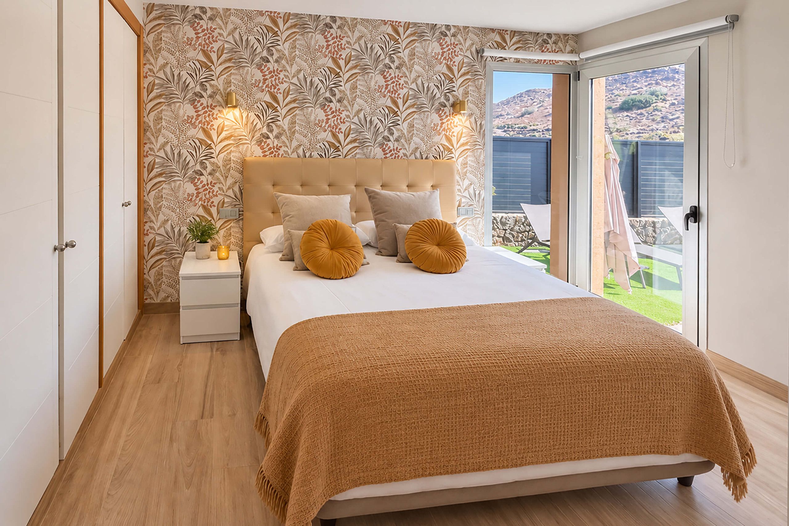 Een slaapkamer met een groot bed, een nachtkastje, een kamerplant en uitzicht op een tuin door een schuifdeur.