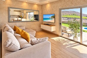 Een moderne woonkamer met een bank, een flatscreen televisie, een groot raam met uitzicht op een strand en een houten vloer.