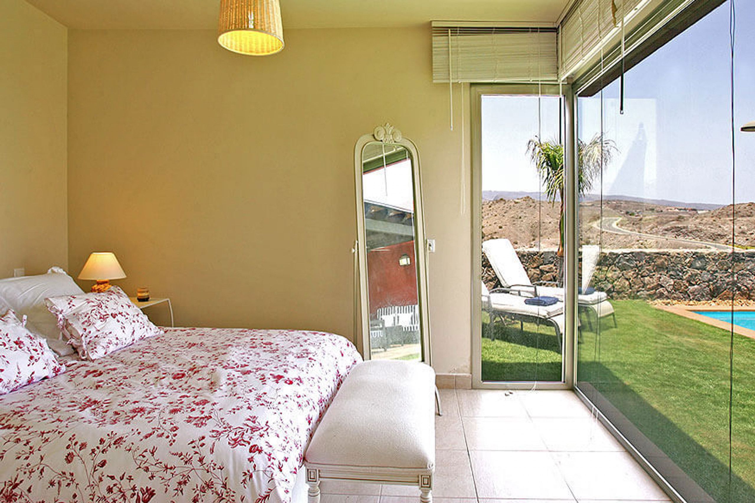 A bedroom with a large bed, a bedside lamp, and a sliding glass door leading to a balcony with a pool view.