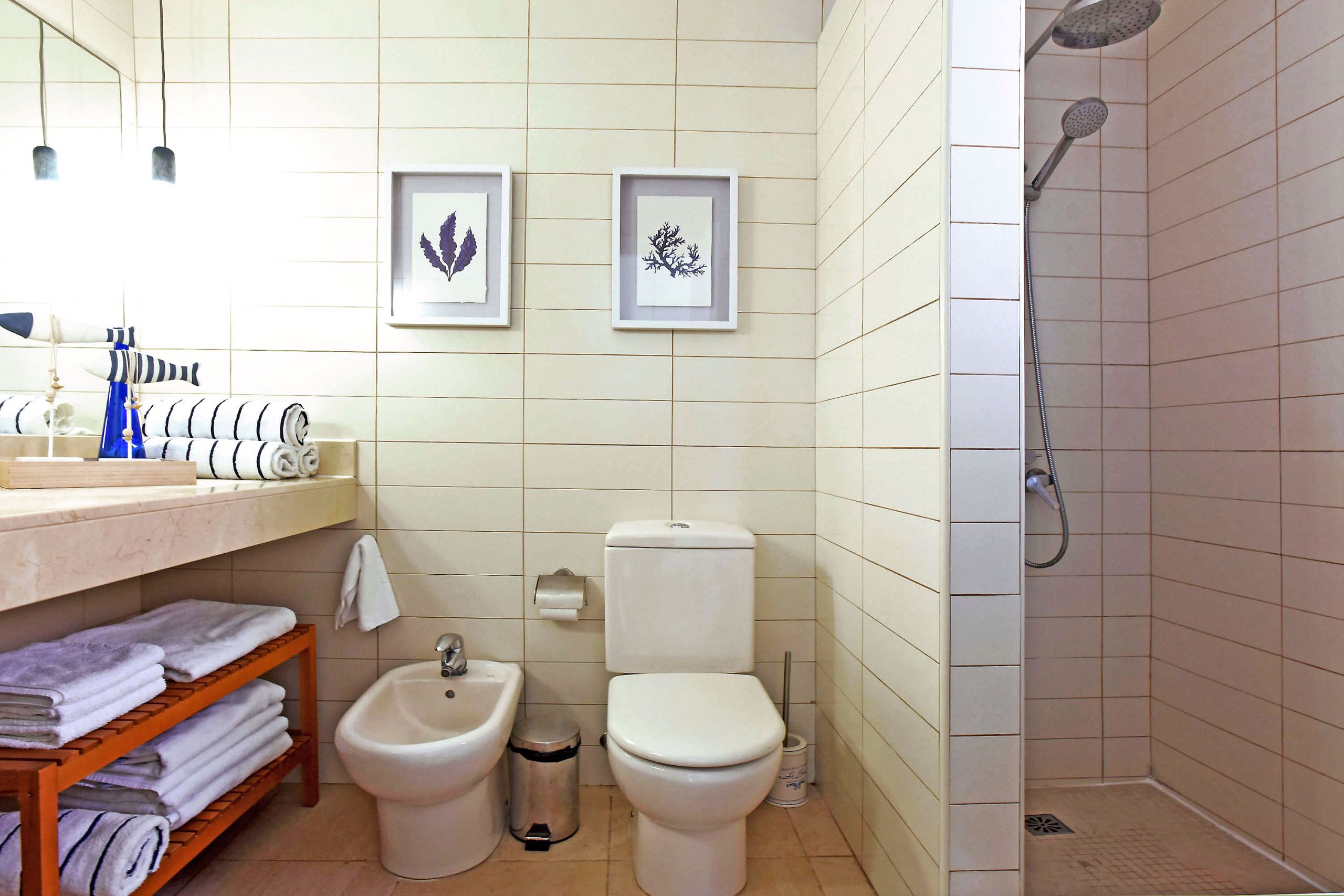 Une salle de bain comprenant des toilettes, un bidet, un lavabo et une douche. Des tableaux encadrés ornent le mur et des serviettes sont posées sur une étagère.