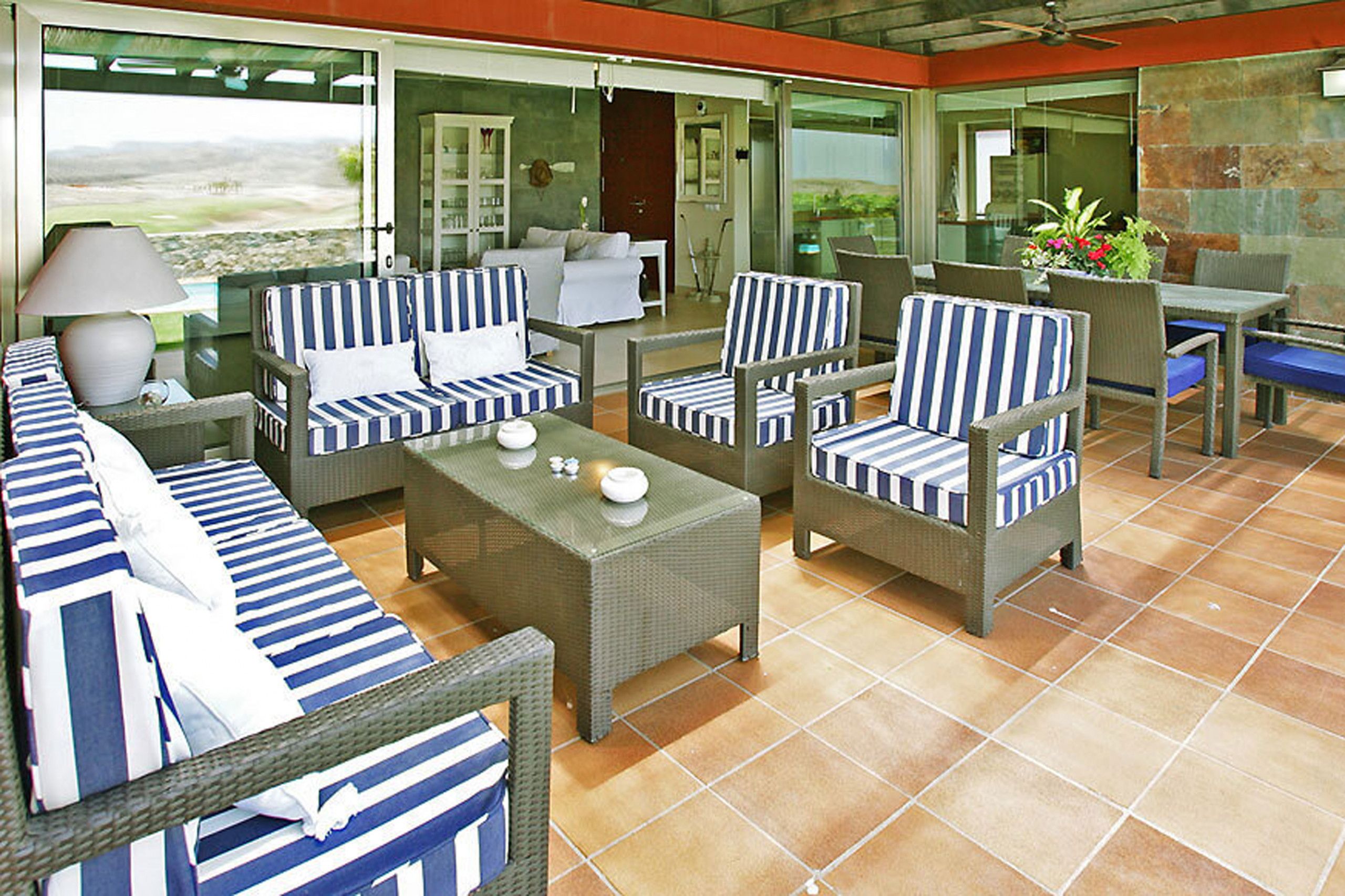 Un patio iluminado por el sol con mobiliario de mimbre, incluyendo sofás y sillones, y una mesa con lámpara.