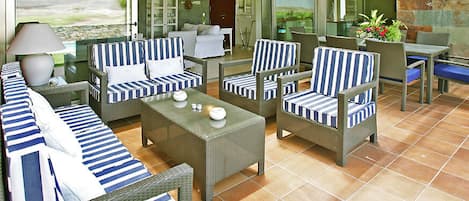 Un patio iluminado por el sol con mobiliario de mimbre, incluyendo sofás y sillones, y una mesa con lámpara.