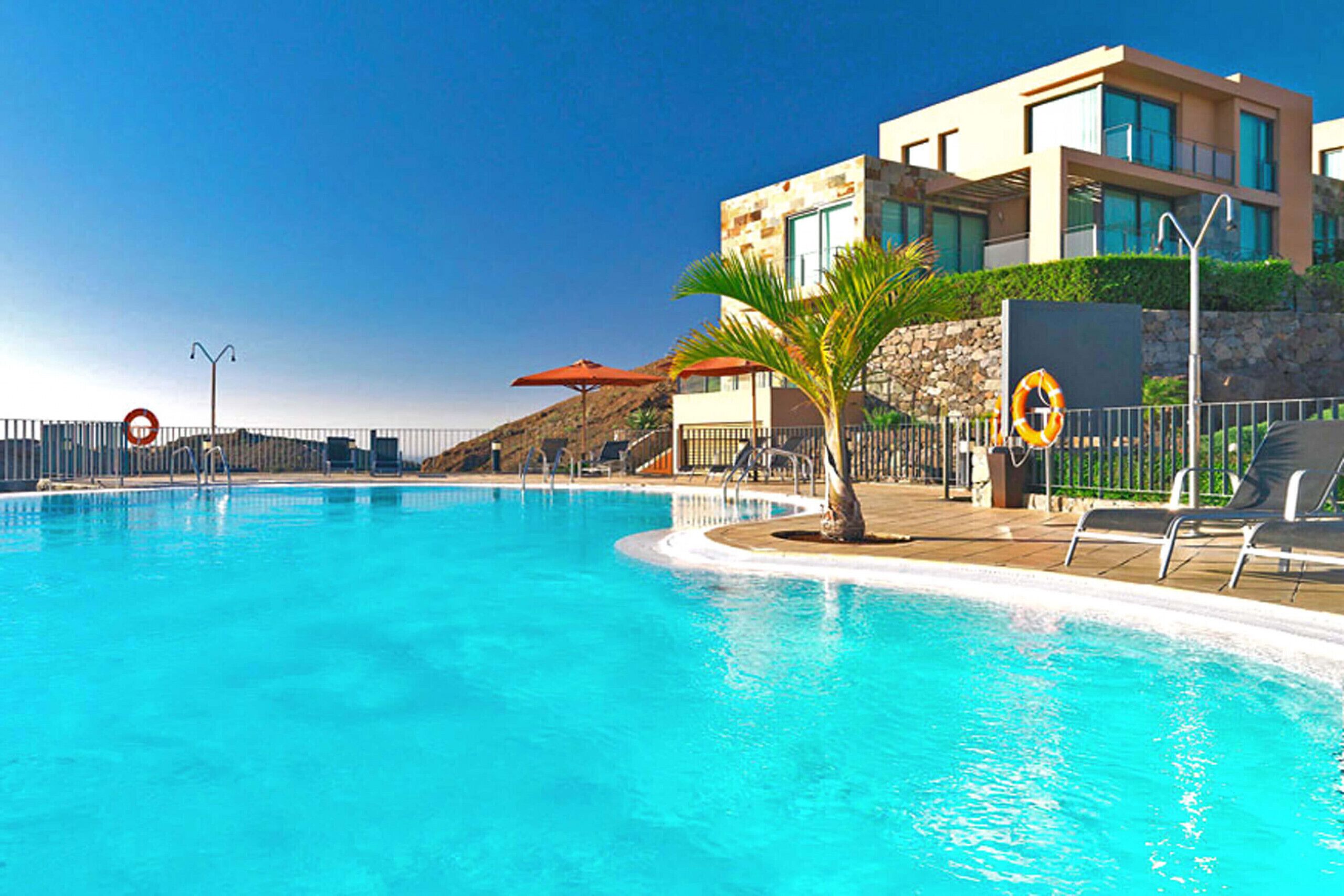 Una piscina con un edificio moderno, palmeras y una silla de salvavidas.