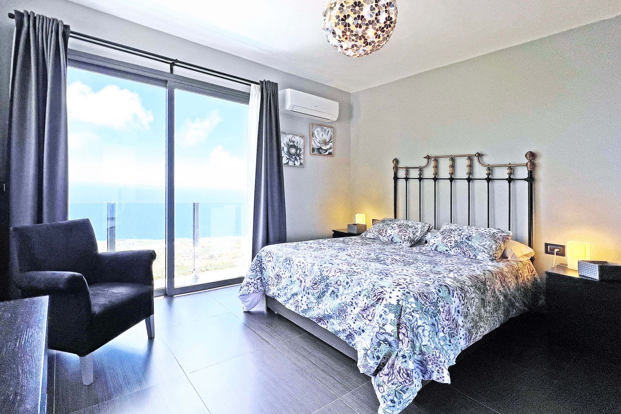 A bedroom with a bed, a chair, a nightstand, and a view of the sea.