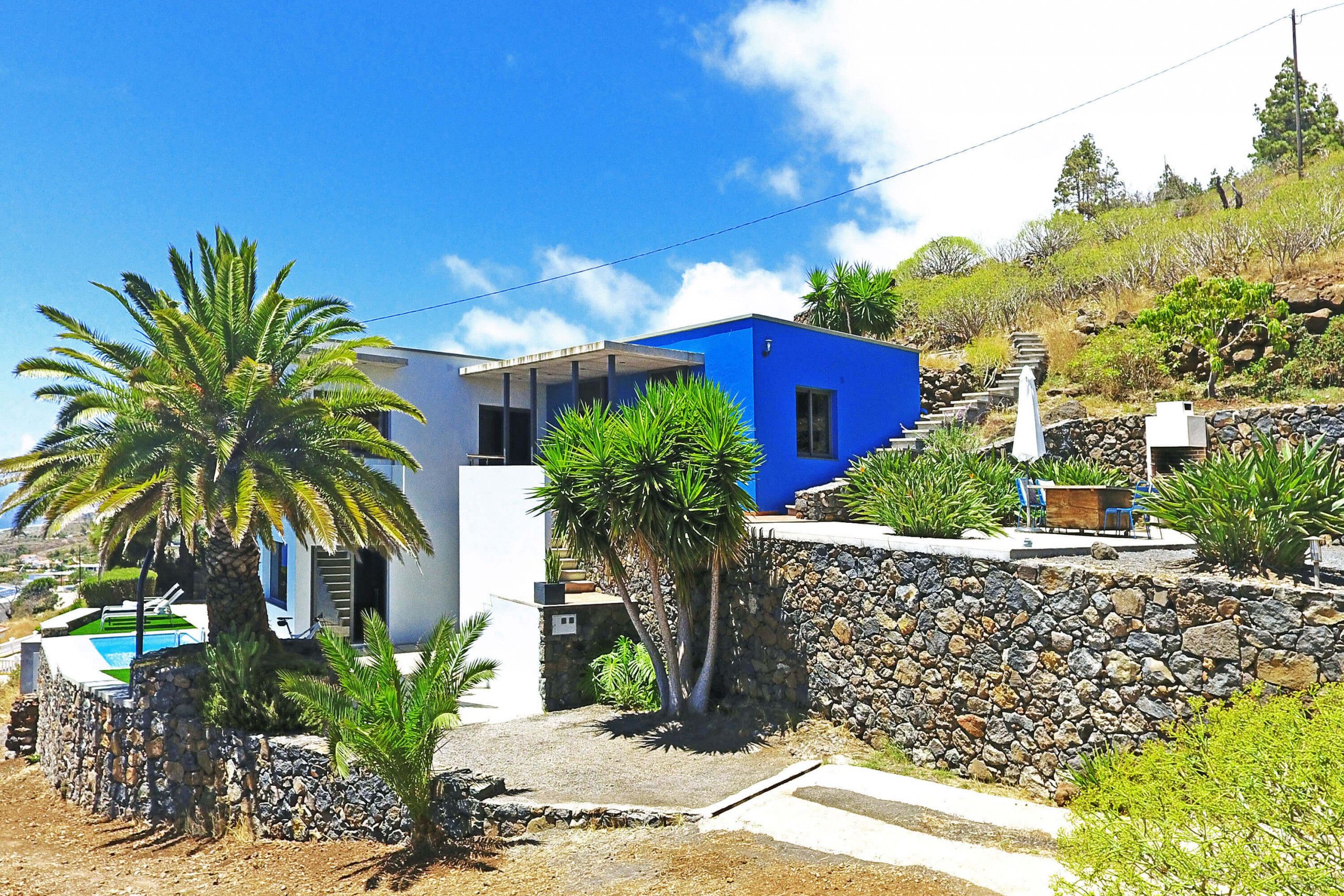 Una casa moderna con fachada azul, un muro de contención de piedra y un área de piscina con palmeras.