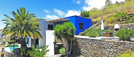 Una casa moderna con fachada azul, un muro de contención de piedra y un área de piscina con palmeras.