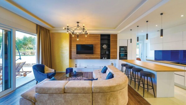 Una sala de estar moderna con un amplio sofá en forma de L, un comedor con taburetes de bar, una cocina con una isla grande y un televisor montado en la pared.